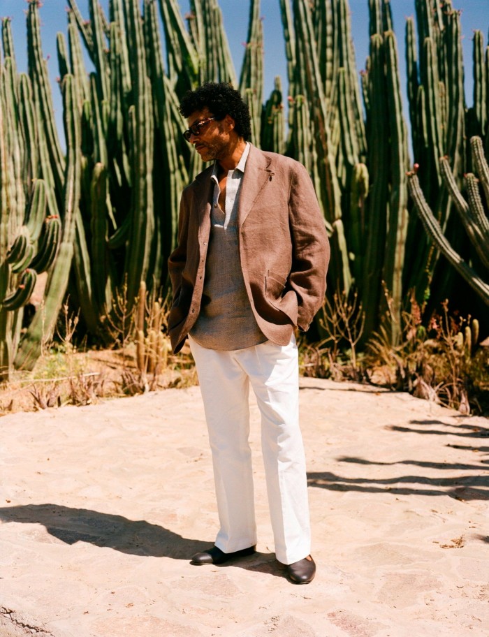 A look from The Anthology’s spring/summer 2024 campaign: a man wearing a brown linen jacket, white trousers and a grey top standing in front of dozens of tall cacti