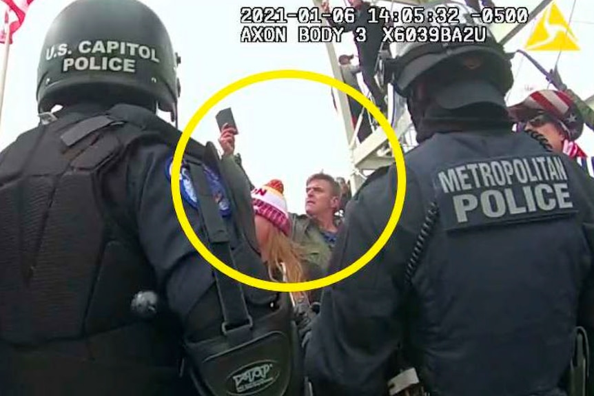 Two metropolitan police with helmets and backs to the camera, a yellow circle around actor Jay Johnston who is holding a phone