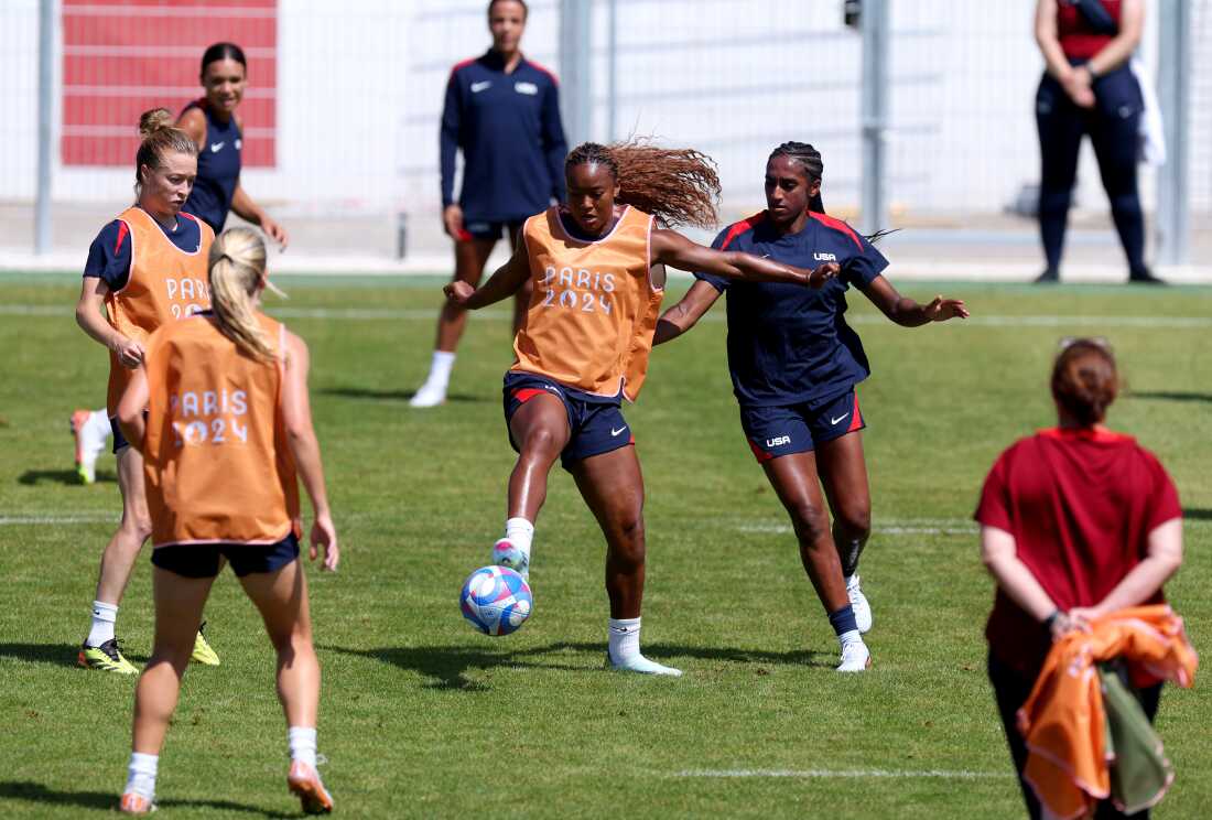 Several women from the U.S. Women's National Team practice soccer on a green soccer field. A few are wearing orange vests that say 