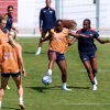 Several women from the U.S. Women's National Team practice soccer on a green soccer field. A few are wearing orange vests that say 