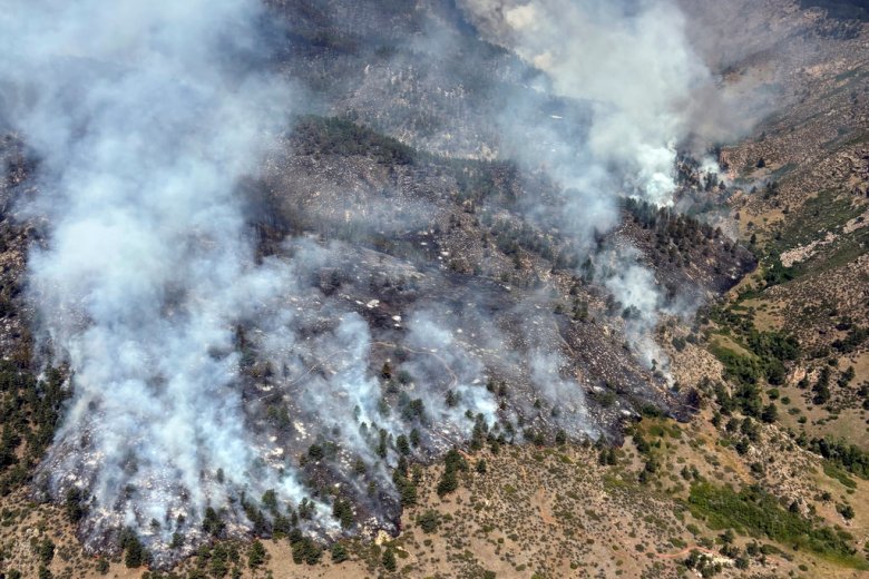 Aerial view of smoke rising from a forest wildfire spreading through a mountainous area, with patches of green vegetation visible amidst burnt sections.