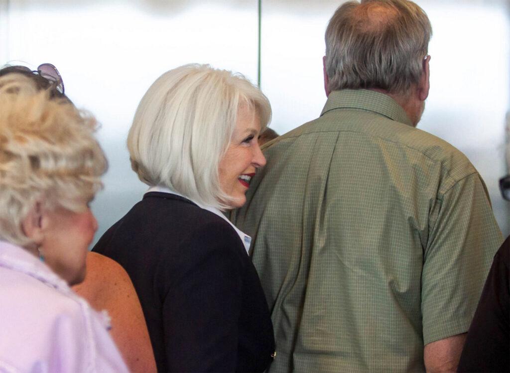 Tina Peters, seen in profile with white hair and a blue blazer, stands in a crowd of people in front of elevator doors.