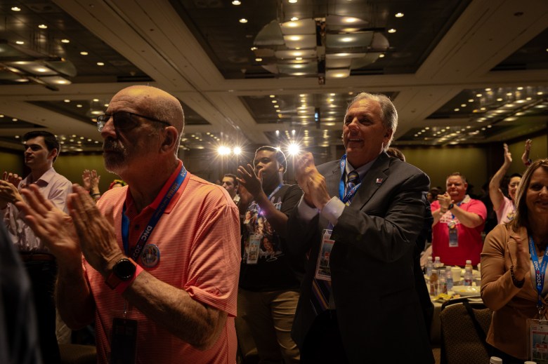 Four warm-colored spotlights shine through crowded venue hall as attendees stand-up and clap during a California Democratic Party delegation breakfast event.