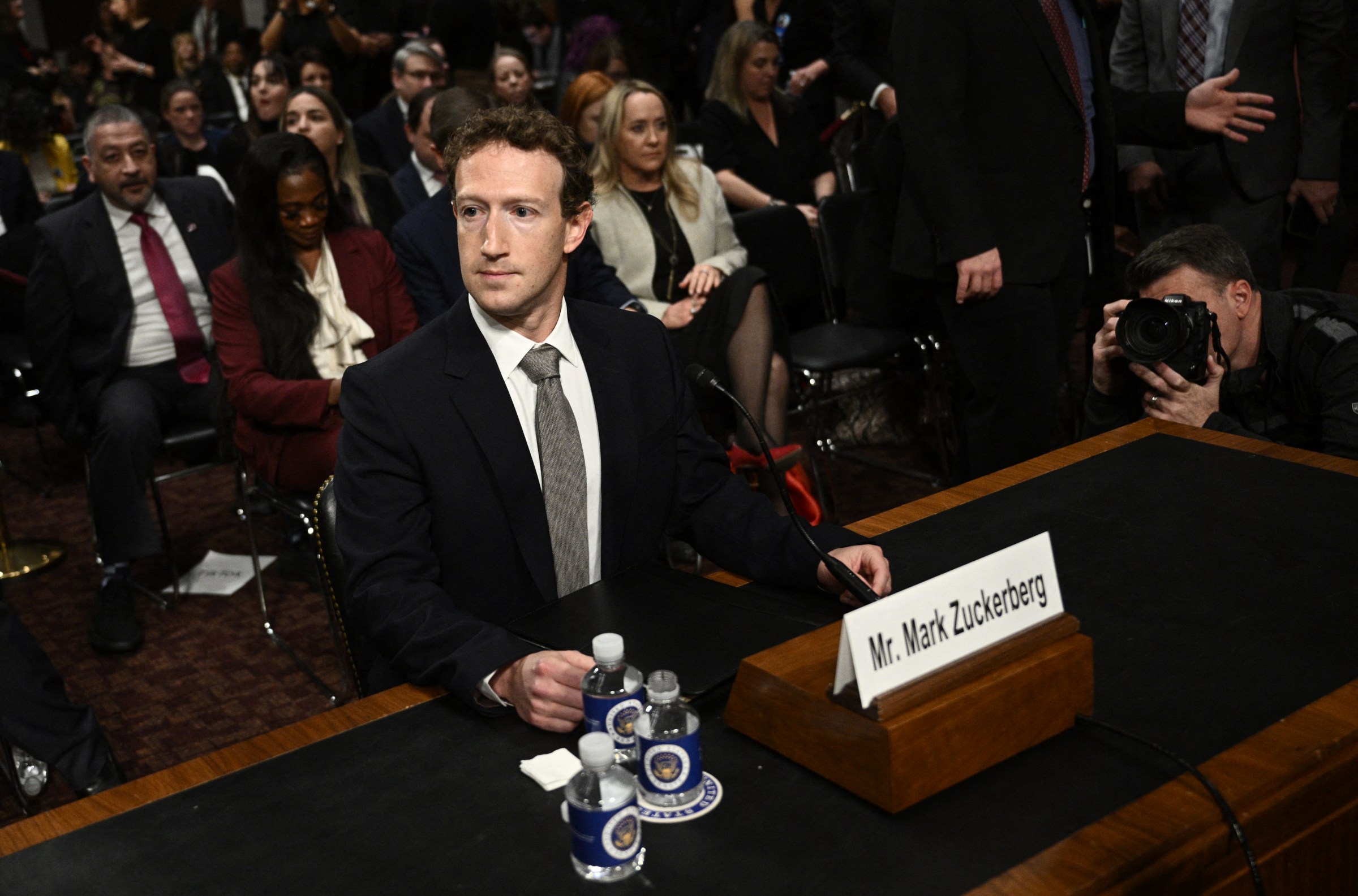 Mark Zuckerberg, in a black suit and gray tie, sits behind a nameplate with his name on it at a Senate desk.