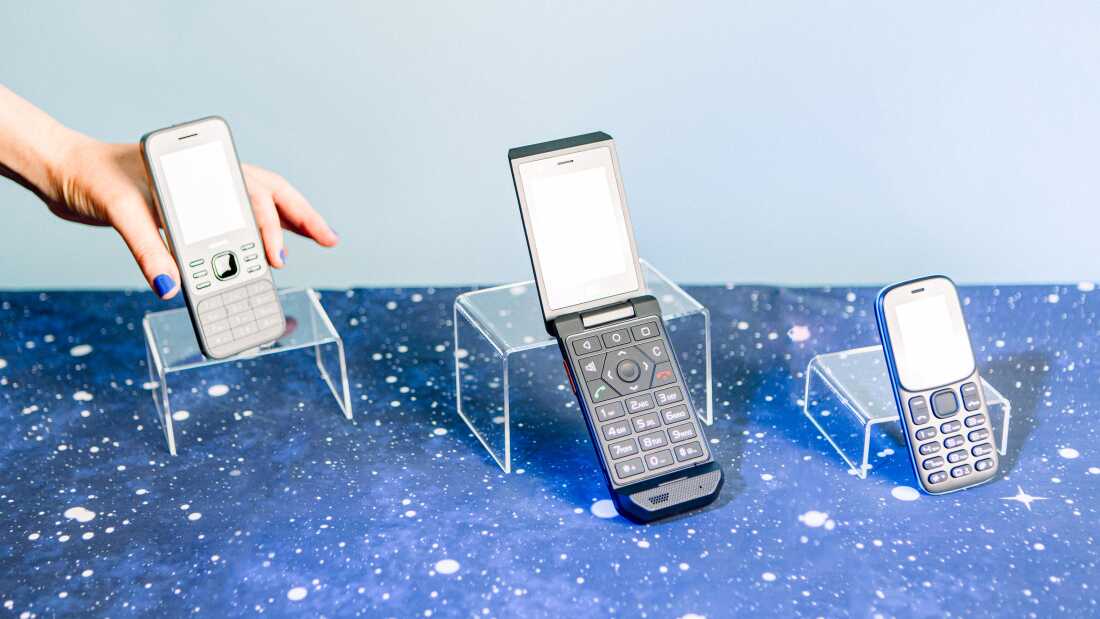 A studio photograph of three basic phones, the Nokia 6300 4G, the Cat S22 Flip and the BLU Z5 -GSM, propped up on clear display shelves. They sit on a table covered with celestial-patterned cloth and behind them is a light blue backdrop. A white hand with blue finger nails reaches in from the top left corner to take the Nokia 6300 4G phone. 