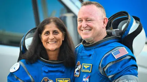 AFP Sunita Williams and Butch Wilmore stand smiling for the camera while wearing astronaut suits