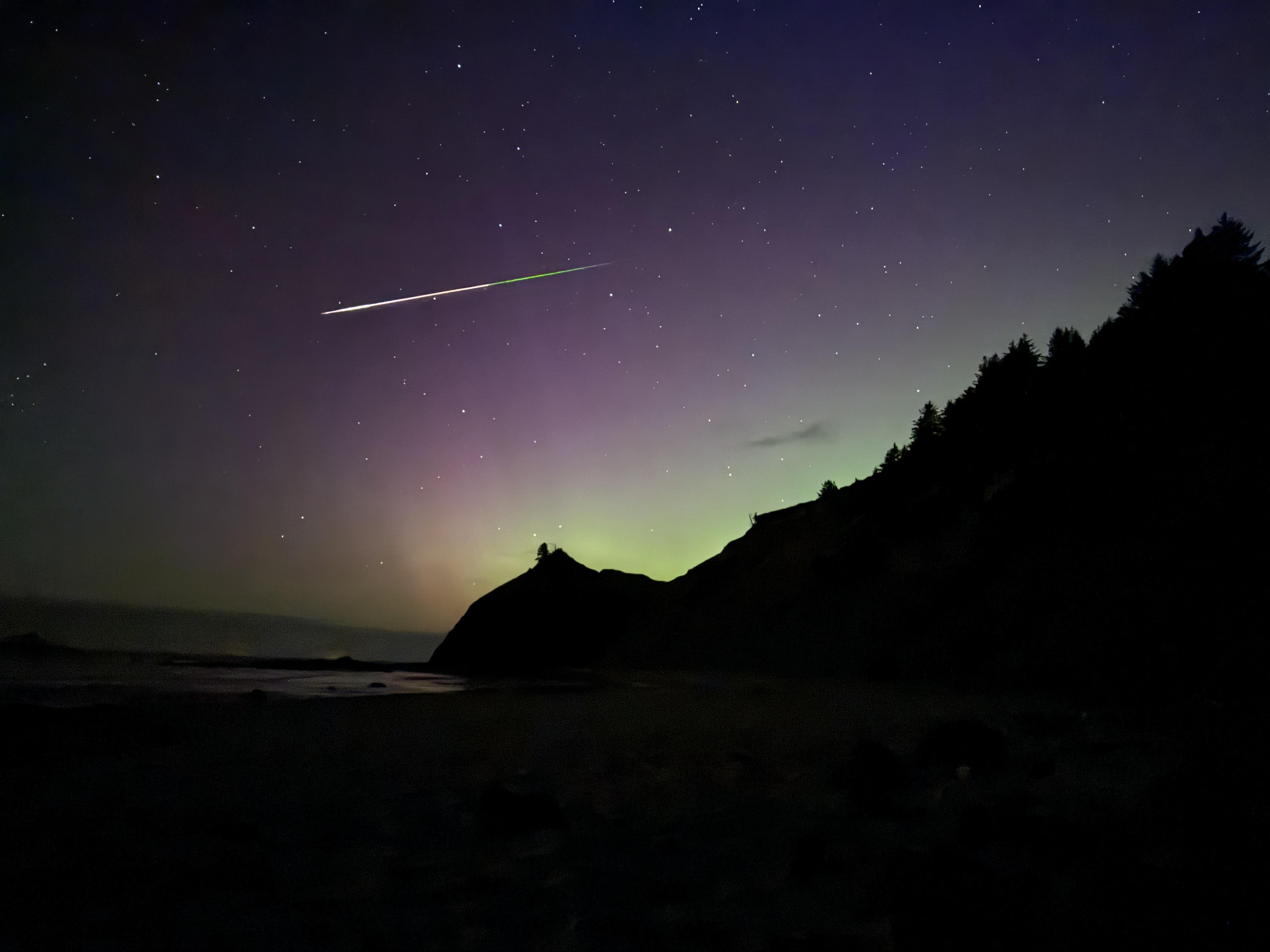 a long colorful meteor streak through the sky with a distinct green glow toward the end of the tail meanwhile the sky glows green and purple during the aurora display.