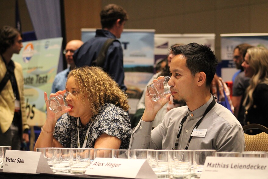 A man sips from a glass of water while seated behind a table full of drinking glasses