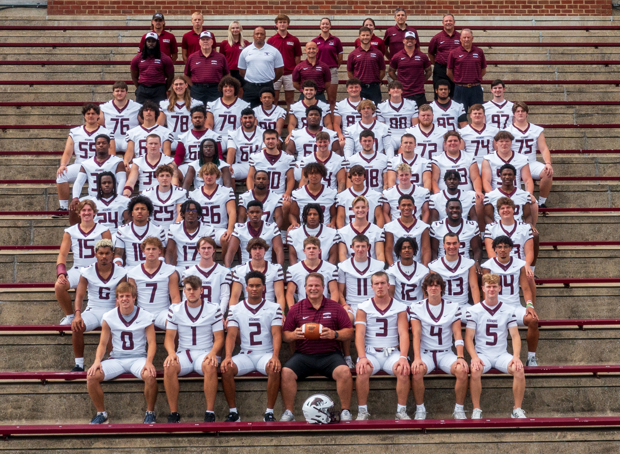 The Roanoke College football team, wearing white uniforms with maroon trim, poses for a team photo on the bleachers at Salem Stadium.