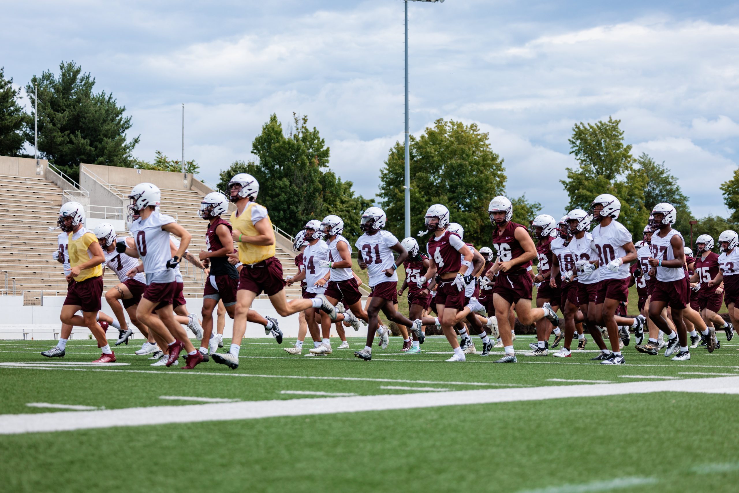 The Roanoke College football team, in white and maroon uniforms, runs on the football field during practice.