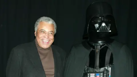 Getty Images A smiling James Earl Jones poses next the right of a person dressed up in a Darth Vader costume during a premiere in New York for Star Wars: Episode II Attack of the Clones in 2002. Jones is wearing a brown sweater with a black jacket over it.