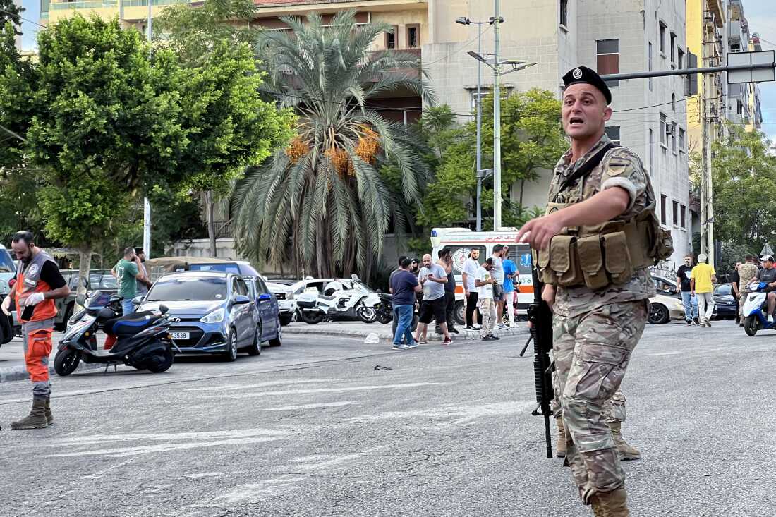 A Lebanese army soldier gestures with one hand at the scene of a reported pager explosion in Sidon, in southern Lebanon, on September 18, 2024. Standing in the street, the soldier is wearing a camouflage-patterned uniform and is holding a firearm. People are standing a short distance behind him. Cars, buildings and trees also appear in the background.