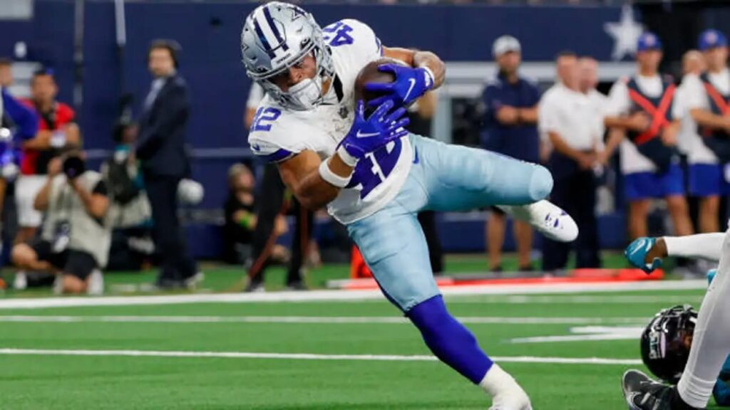 Cowboys football player Deuce Vaughn in blue and white uniform running with the ball, with defenders attempting to tackle him on the field.