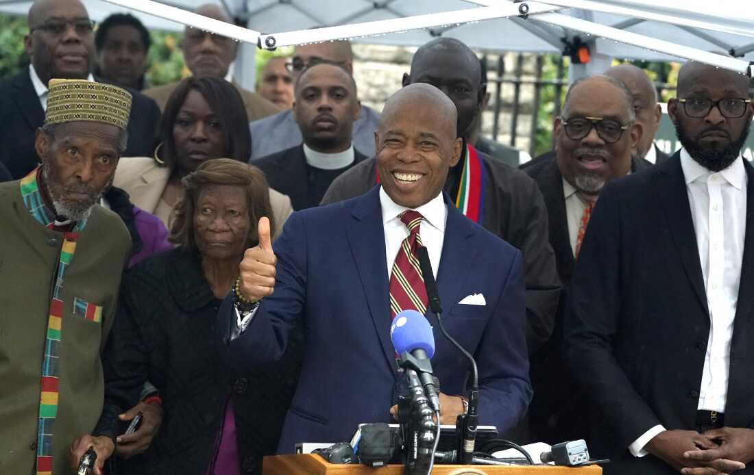 In this photo, New York City Mayor Eric Adams holds a news conference outdoors on September 26, 2024. Supporters are standing behind him, and Adams, wearing a suit and tie, stands at a lectern with microphones attached to it.