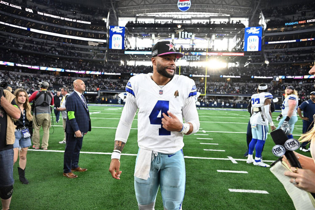 Football player in a white jersey with number 4 walks on the field, smiling, while surrounded by people and press in a stadium.