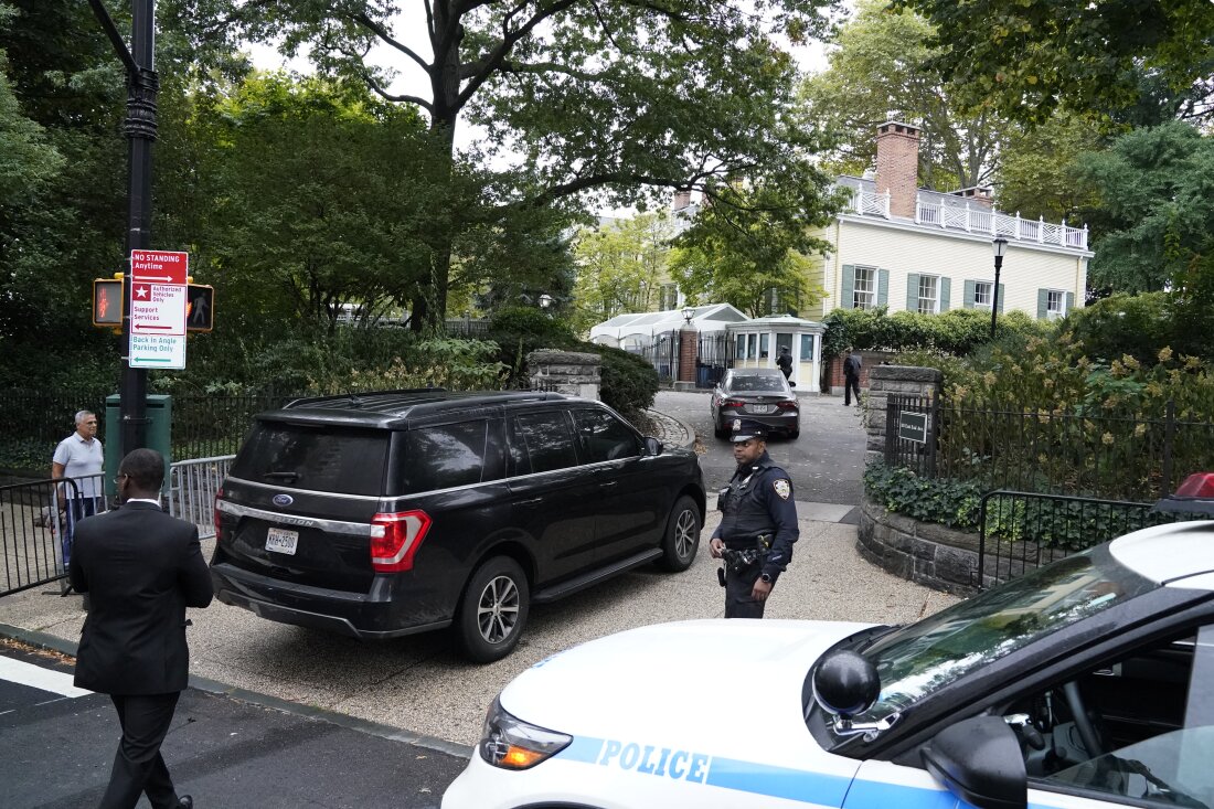 In this photo, a dark SUV enters the driveway of Gracie Mansion, the official residence of New York City's mayor, on September 26, 2024. A police officer stands to the right of the SUV, and a police car is parked on the side of the road in front of Gracie Mansion. The butter-colored home stands in the background, surrounded by trees.