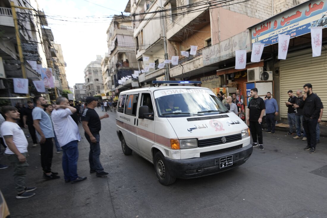 In this photo, an ambulance believed to be carrying wounded people drives down a street in a southern suburb of Beirut on September 18, 2024. The ambulance is white with a blue light on top that spans the width of the vehicle. Men stand on the sides of the street, which is lined with buildings.