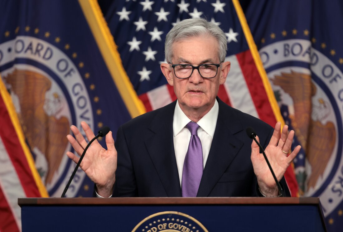 This photo shows Federal Reserve Chair Jerome Powell standing behind a lectern that has two microphones. He's wearing a suit and glasses. American flags and also flags with the Federal Reserve seal are in the background.