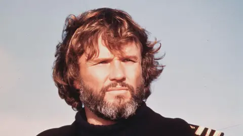 Getty Images Kris Kristofferson staring off into the distance, squinting a little like the sun is in his eyes. He has shaggy brown hair, a greying beard and the sky is light blue behind him.