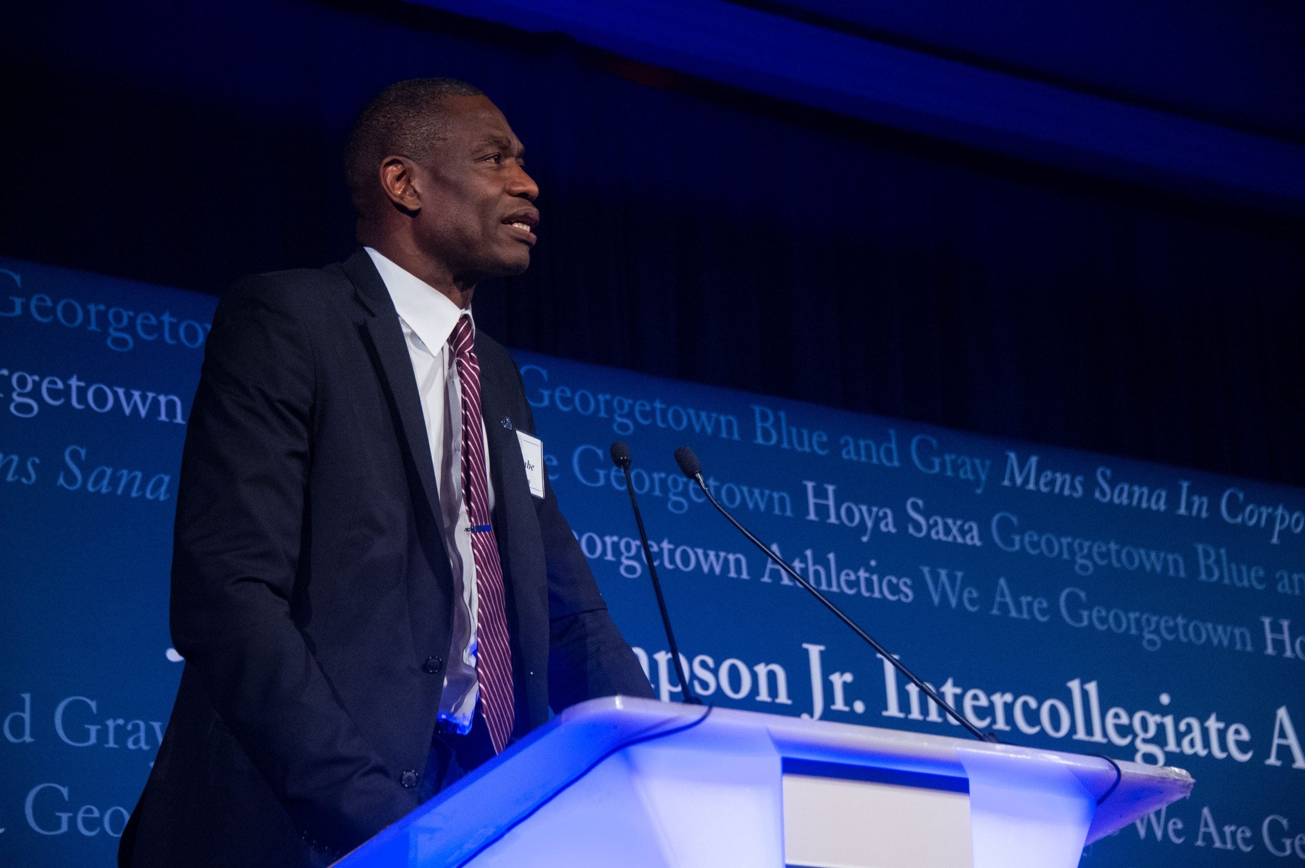 Dikembe Mutombo in a suit and tie speaking at a podium