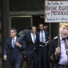 This photo shows two attorneys for former President Donald Trump — John Lauro (on the far left) and Todd Blanche (second from left), departing through double doors at the E. Barrett Prettyman U.S. Courthouse in Washington, D.C., in 2023. A protester wearing a costume in Trump's likeness stands in the foreground, carrying a sign that says: 