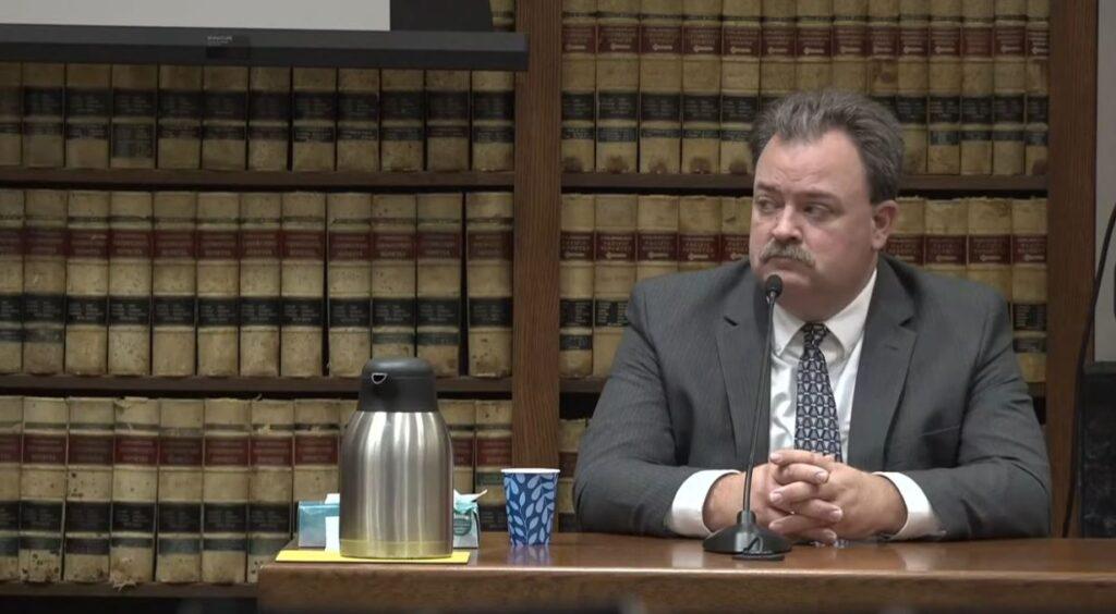 A man named Dallas Schroeder in a gray suit, white dress shirt and triangle-patterned tie sits at a wooden table with a microphone in front of him waiting to testify in a courtroom in Grand Junction.