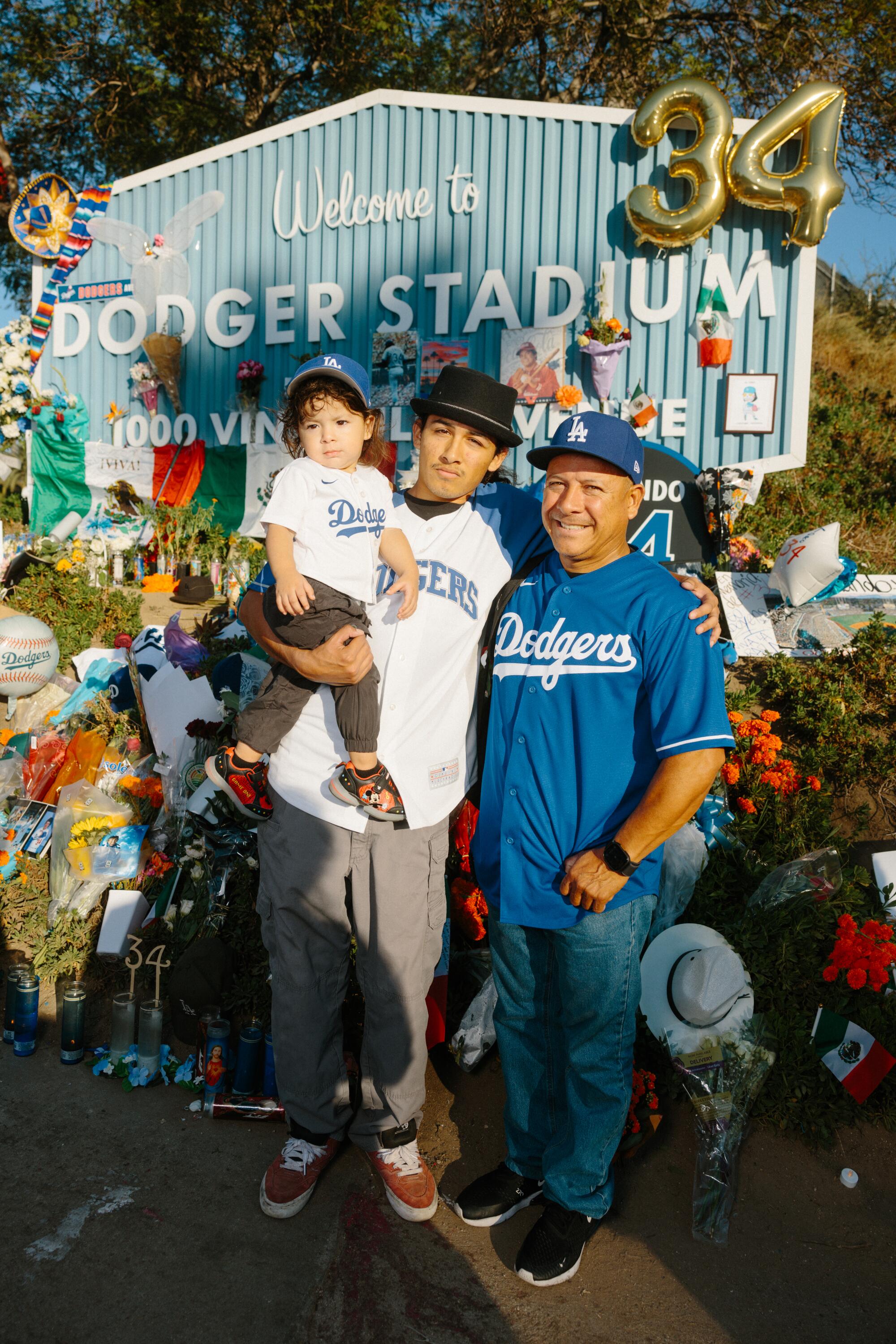 One adult holds a small child and stands next to another adult, all of them in Dodgers jerseys in front of flowers.