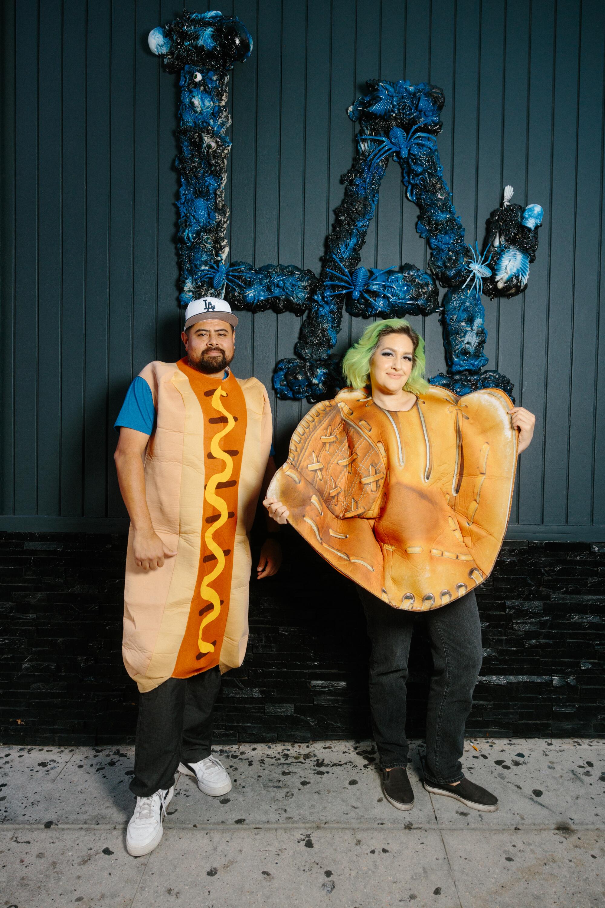 Ralph Gomez and Kristen Hagen stand in their hot dog and glove costumes under giant blue LA letters.