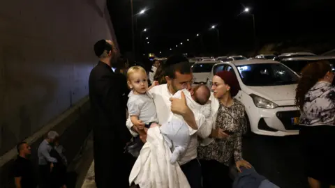 Reuters A man holds children as people take cover as air raid sirens sound across Israel during an Iranian ballistic missile attack, in central Israel (1 October 2024)