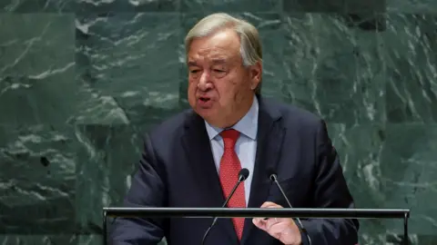 Reuters UN Secretary General Antonio Guterres giving a speech at the United Nations in New York. He is standing against the distinctive green marbled backdrop at a lectern, wearing a red tie, dark suit jacket and blue shirt