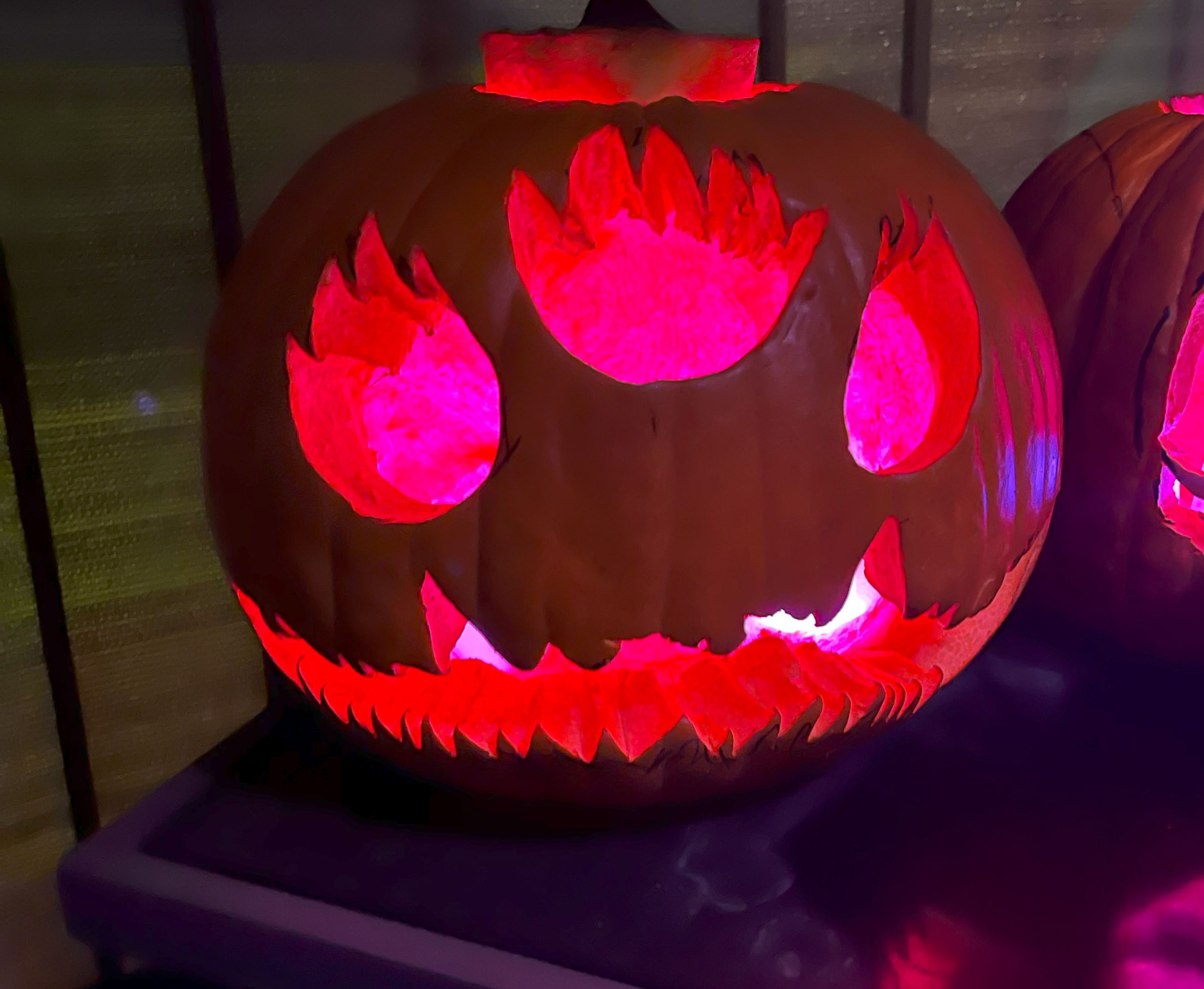 A jack-o-lantern with three eyes that look like flames, and a jagged long mouth. Its features are lit with pink lights.