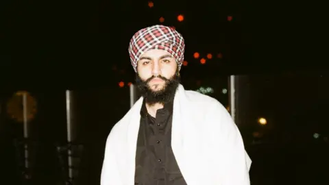 Beant Dhillon Beant wears a red and white checkered turban with a black top and white shawl.