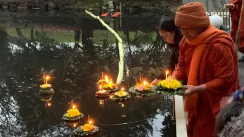 Venerable Phramaha Bhatsakorn Piyobhaso The Venerable wears orange drapes and an orange hat, holding a krathong with green banana leaves and orange flowers and a candle in the middle. He is releasing it into a large pond of water. 