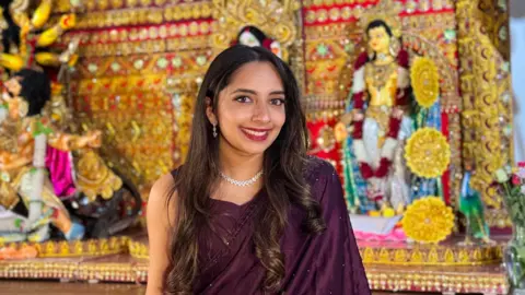 Vedika Apte Vedika is dressed in a dark purple saree wearing her hair in loose curls and smiling. She stands in front of a Hindu shrine.