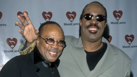 Getty Images Quincy Jones and Stevie Wonder standing together and smiling on an awards ceremony red carpet in 1999