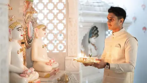 Anil Photography Dev standing on the right wearing a cream coloured kurta or traditional Indian top. He is praying while holding a plate with offerings and a diva in front of two white statues.